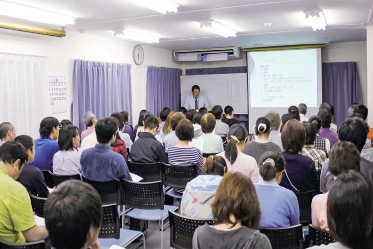 介護施設での講演会やレクリエーション。大勢の入居者が集まりプロジェクターを使った説明を受けている。